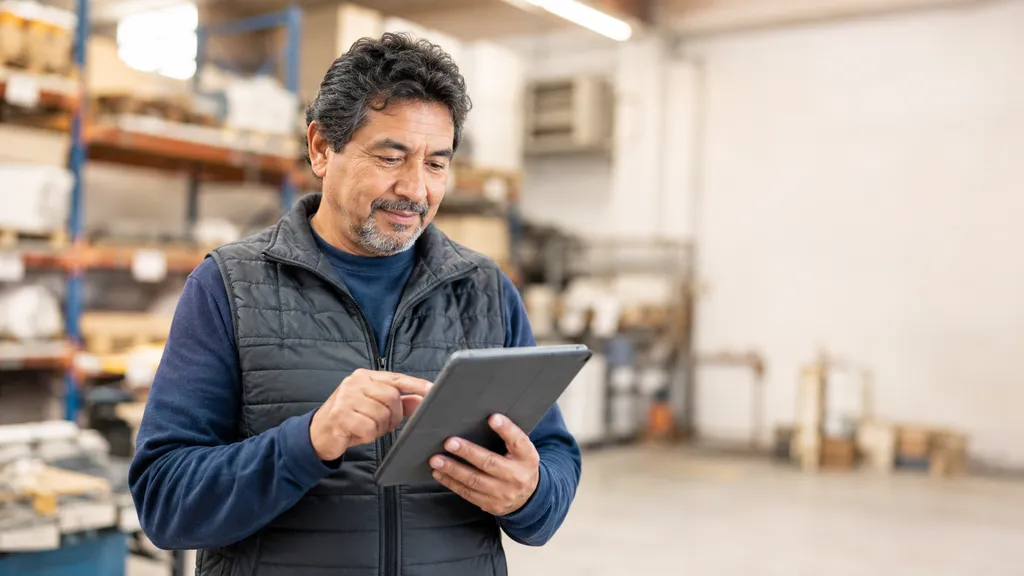 A man stands in a warehouse, using a tablet while wearing a gray vest and blue shirt. Shelves with boxes and equipment are in the background.